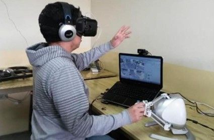 An operator with a VR headset and headphones controls a haptic device next to a laptop
